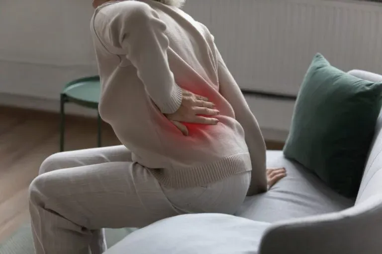 A person in a beige sweater sits on a couch, hand on their lower back, indicating back pain. Limitless Spine & Joint Care offers back pain relief in Morganville, NJ.