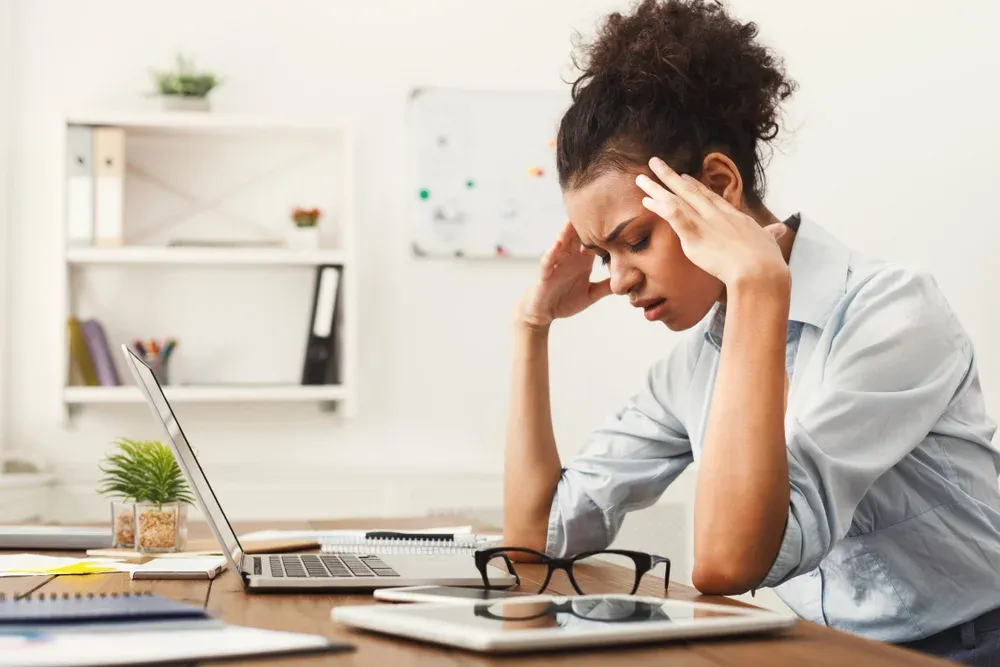 A woman sits at a desk with a laptop, glasses, and tablet, holding her head in her hands, possibly experiencing headaches migraines. Image used by Limitless Spine & Joint Care.